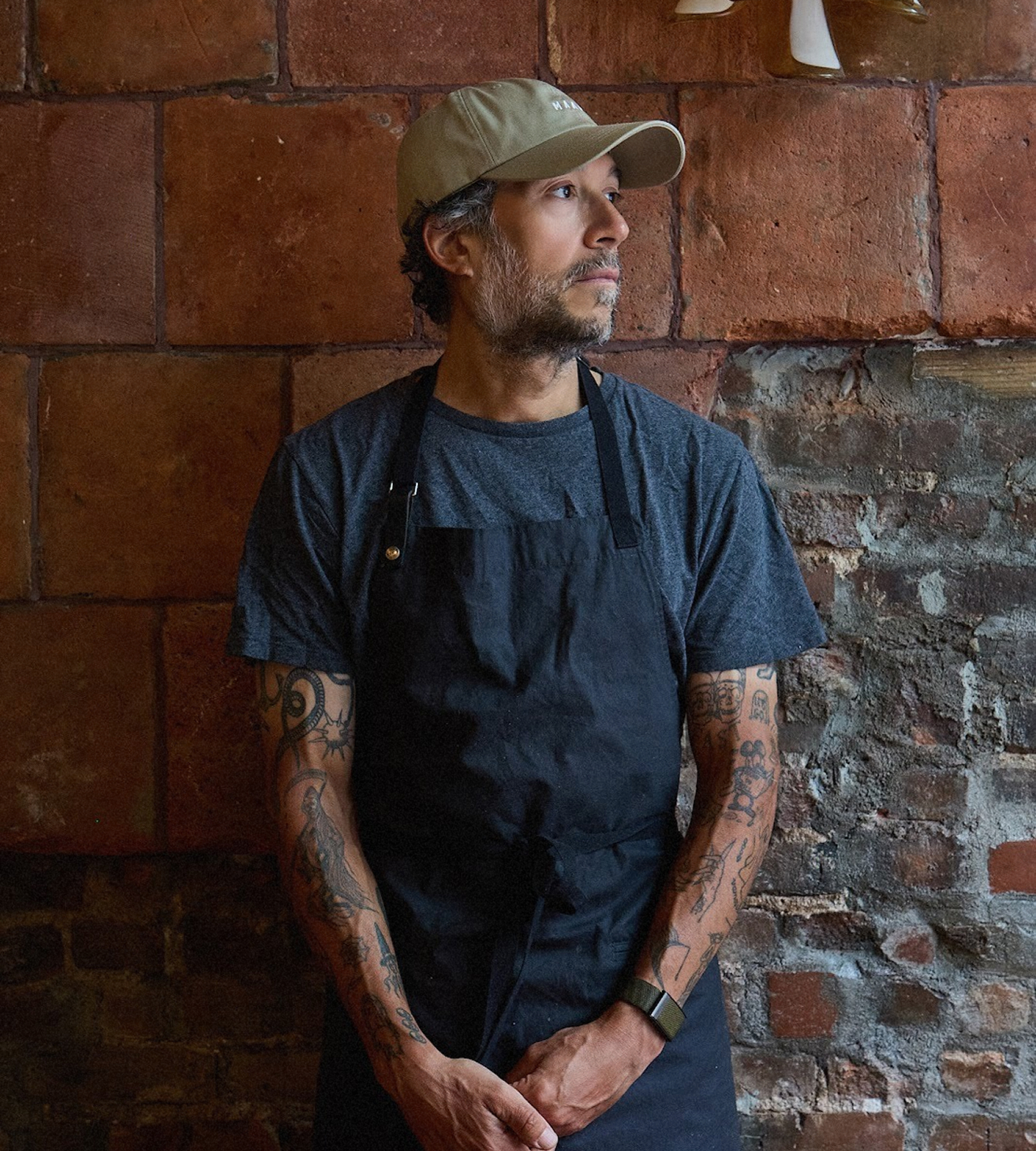 Profile portrait of a tattooed chef wearing a blue apron and beige baseball cap, standing against a rustic brick wall in a restaurant kitchen setting.