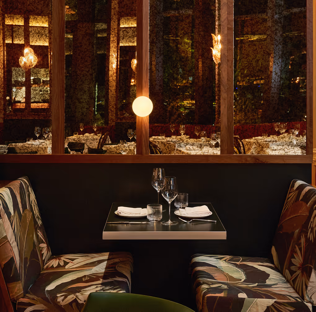 Intimate restaurant table setting with patterned booth seating, wine glasses, and warm lighting behind a wooden partition.