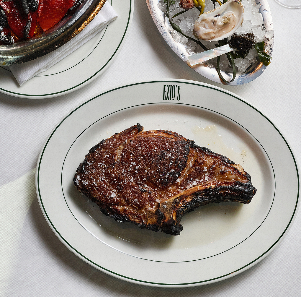 Grilled beef steak served on a white plate with black trim, accompanied by oysters on a white tablecloth.