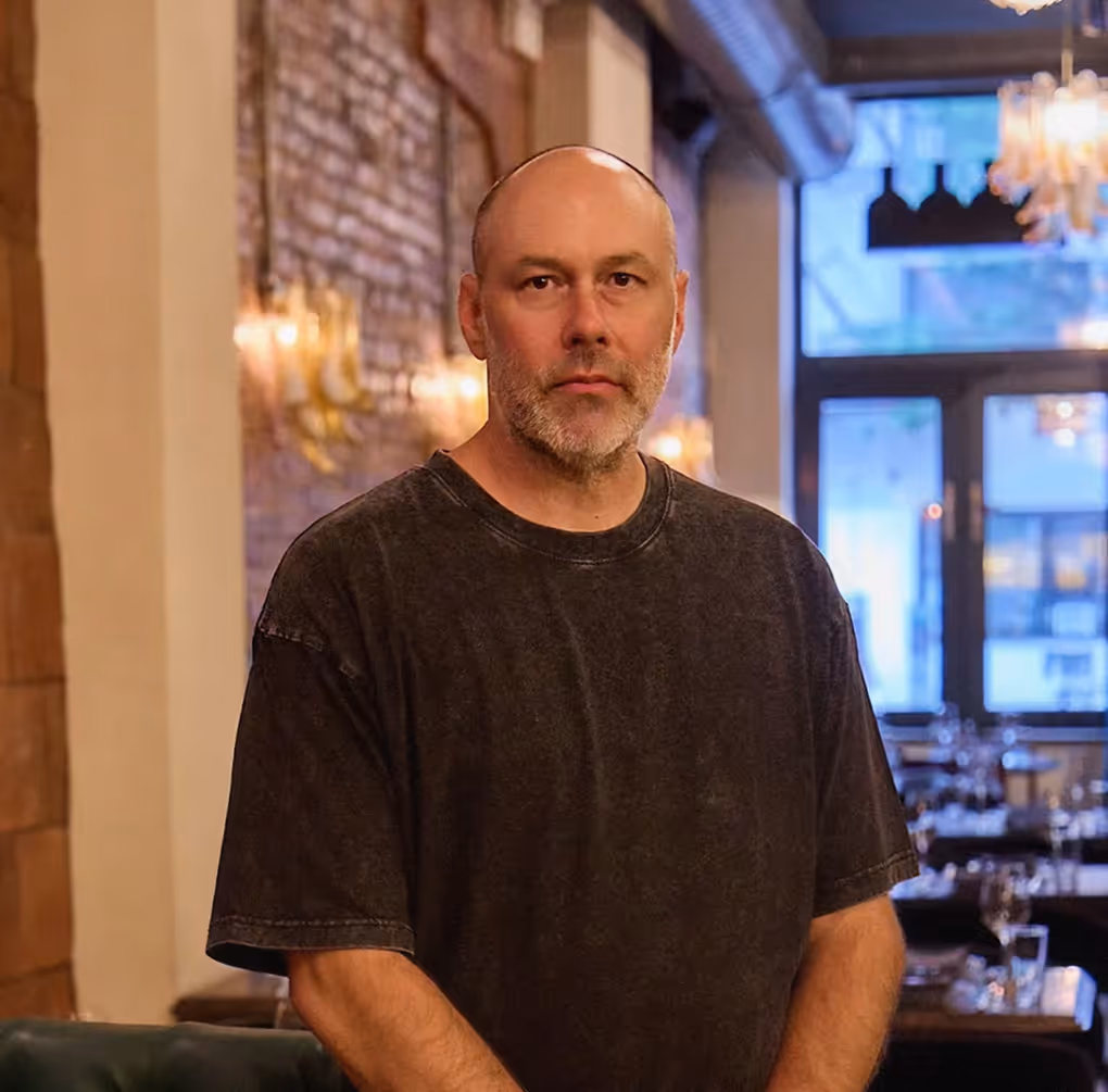 Portrait of a bald man with a short beard wearing a dark grey t-shirt, standing in a modern restaurant with an exposed brick wall and warm ambient lighting in the background.