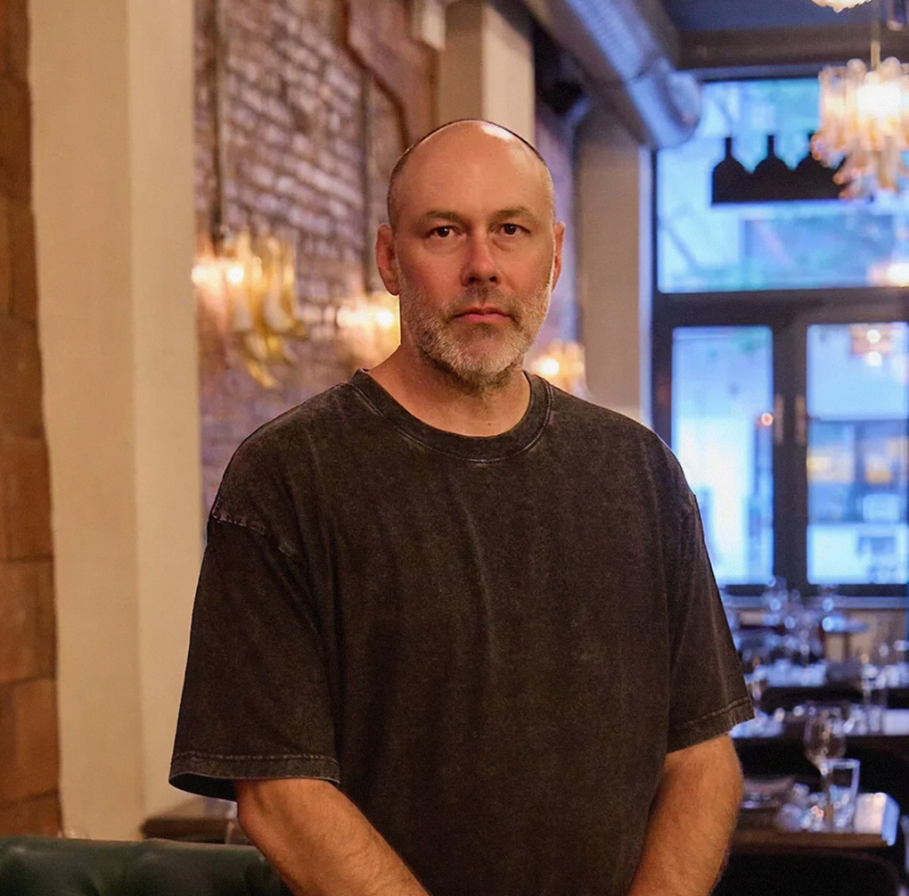 Portrait of a bald man with a short beard wearing a dark grey t-shirt, standing in a modern restaurant with an exposed brick wall and warm ambient lighting in the background.