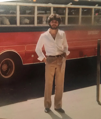 Bearded man wearing a white shirt and beige trousers standing in front of a vintage red bus.