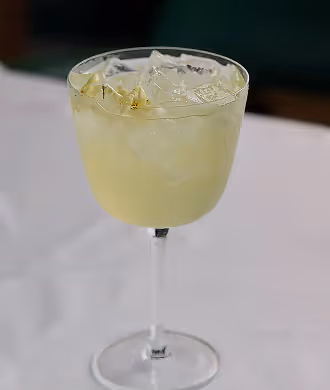 Stemmed glass filled with a pale yellow iced cocktail and lemon zest on a white restaurant tablecloth.