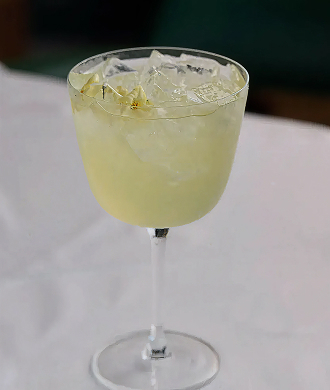 Stemmed glass filled with a pale yellow iced cocktail and lemon zest on a white restaurant tablecloth.
