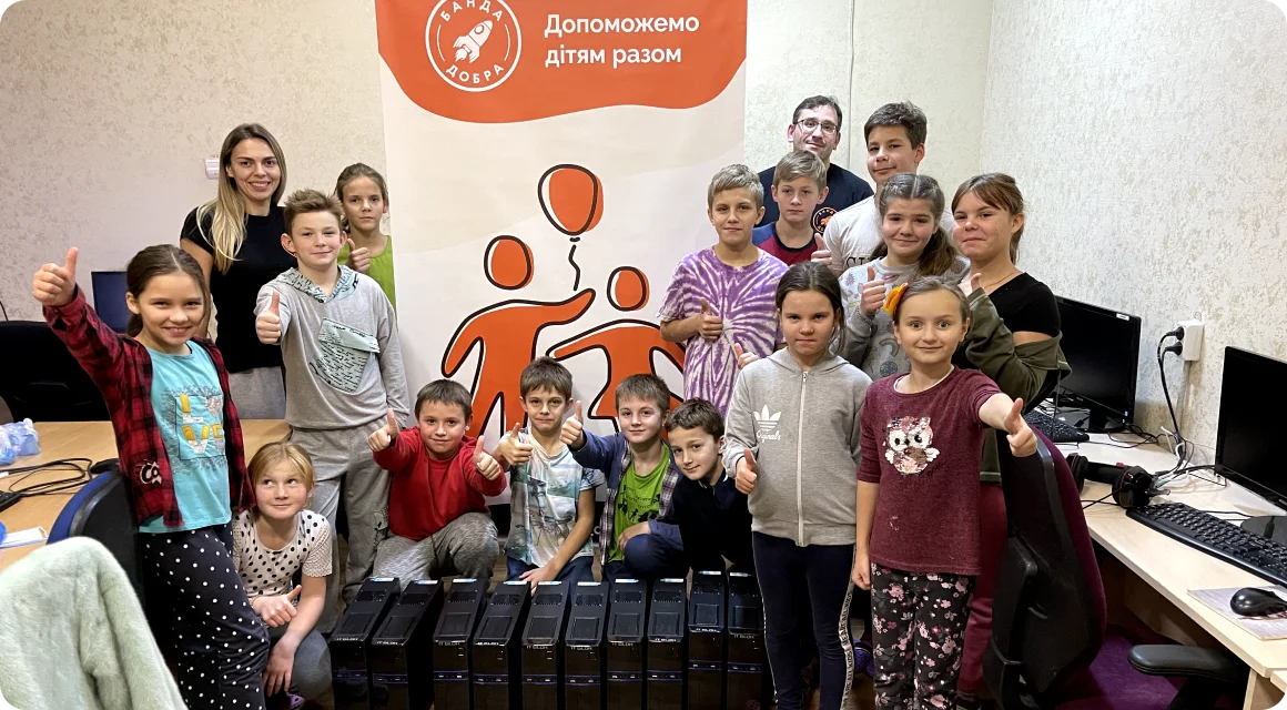 Група дітей та волонтерів Банди Добра у комп’ютерному класі позує з подарованими системними блоками, усі усміхаються та показують “лайк”.