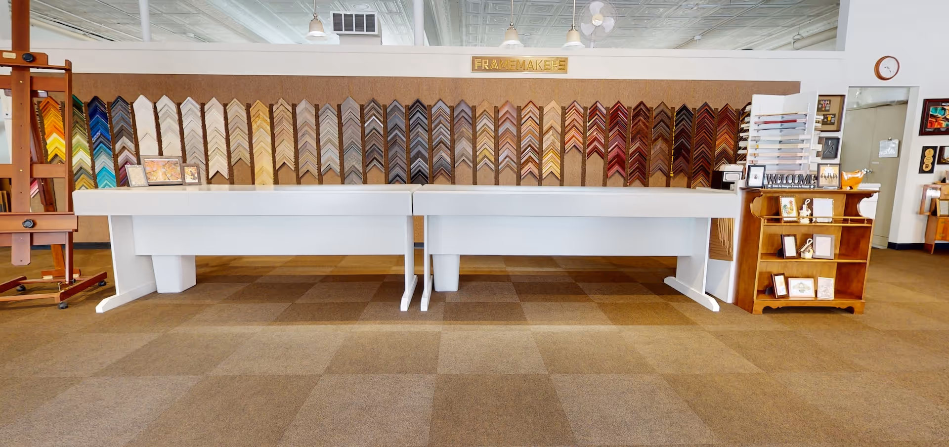 Interior of a frame shop with sample frame corners displayed on a wall behind a white work table and wooden shelving unit.