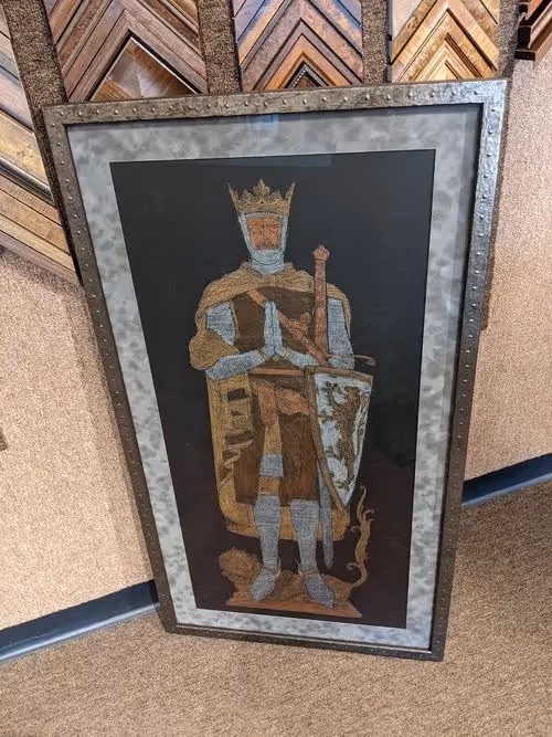 Framed embroidery of a medieval knight or king wearing a crown, holding a sword and shield, with hands clasped in prayer.