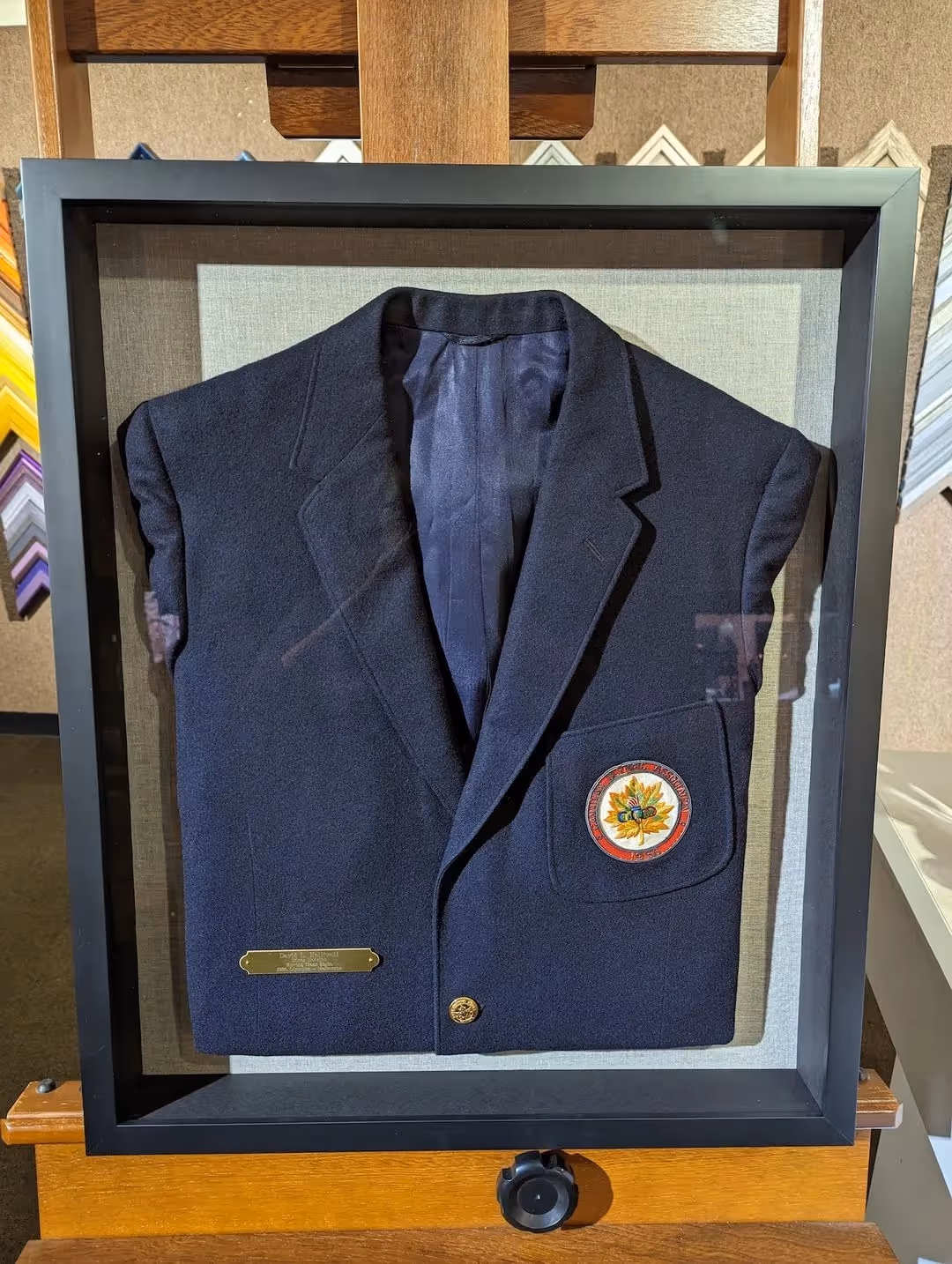 Framed dark navy blue blazer with a patch featuring a maple leaf and other symbols on the left chest pocket, displayed on a wooden easel.