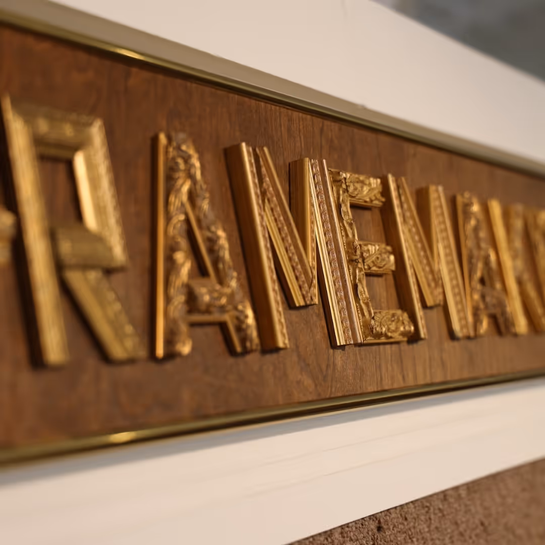 Close-up of ornate gold letters spelling 'RAME' mounted on a wooden board with a gold frame.