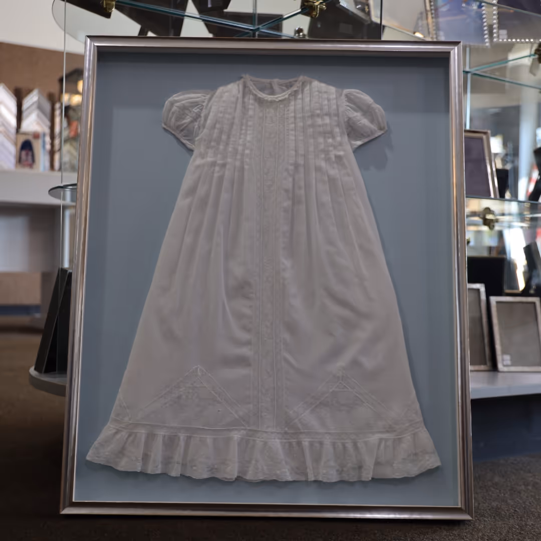 White vintage baby dress with puffed sleeves and lace details, displayed in a silver frame.