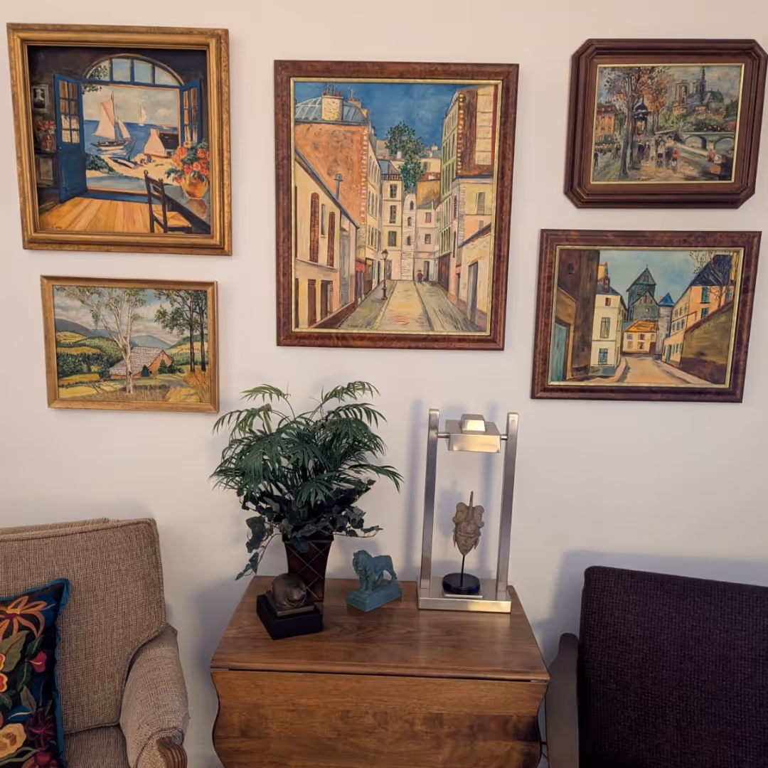 Wall with five framed paintings of landscapes and city scenes above a wooden table holding a potted plant, a lion figurine, a sculpture in a glass case, flanked by two upholstered chairs.