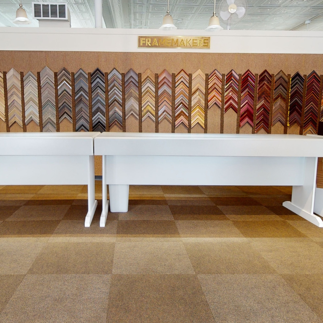 Interior of a framemaker shop with a wall display of various picture frame samples arranged by color and style above white tables.