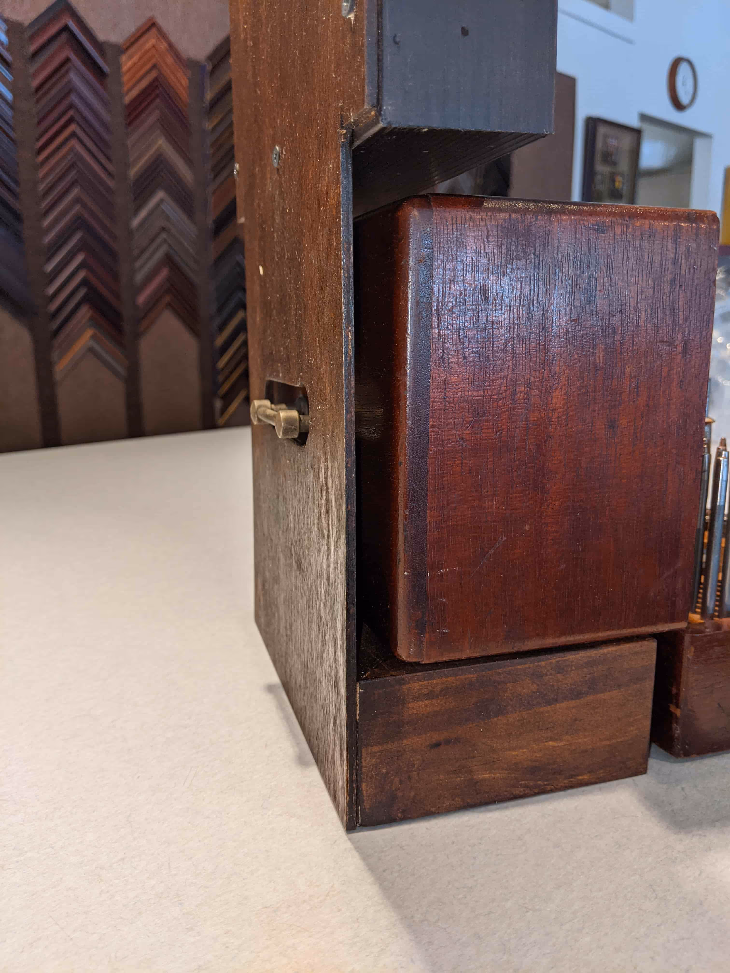 Close-up of a dark wooden box with a brass latch on a light-colored table, with picture frame samples and a clock on the wall in the background.