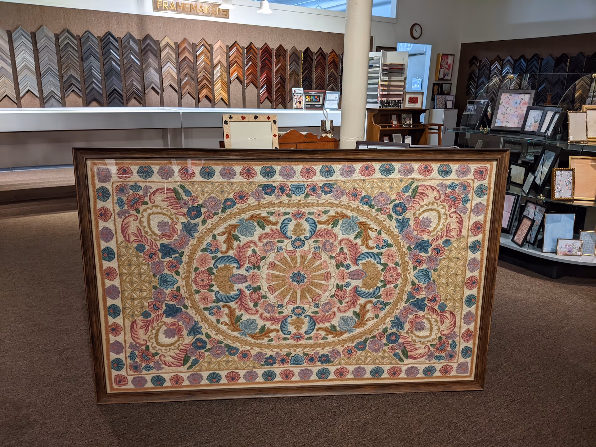 Framed ornate rug with floral and geometric patterns in pastel colors on display in a framing shop.