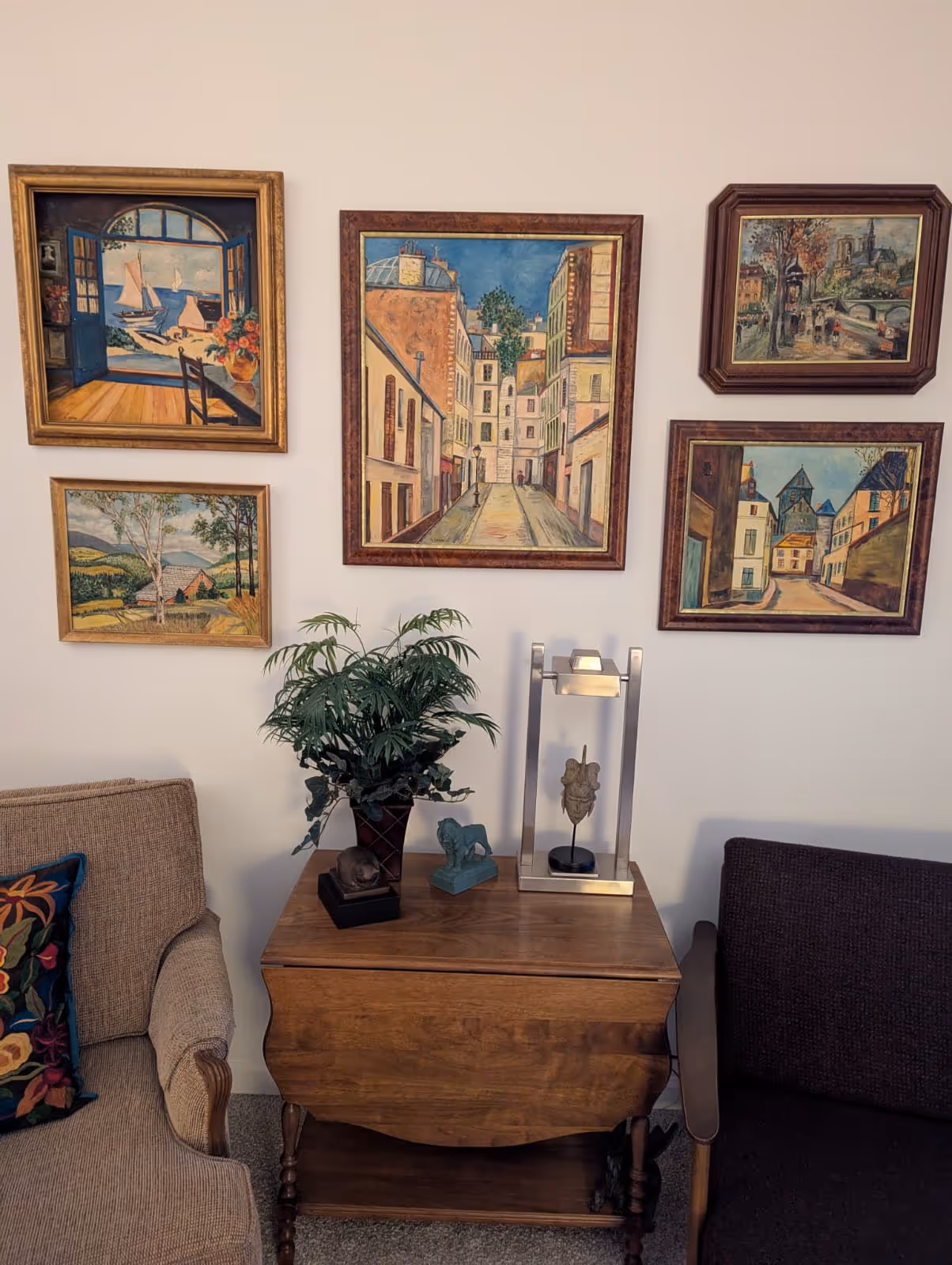Wall with five framed paintings depicting landscapes and cityscapes above a wooden side table holding a potted plant and decorative sculptures, flanked by two armchairs.