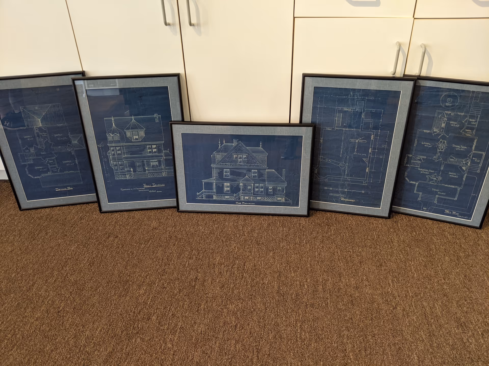 Five framed vintage blueprints of a house showing floor plans and elevations, leaning against white cabinets on a brown carpeted floor.