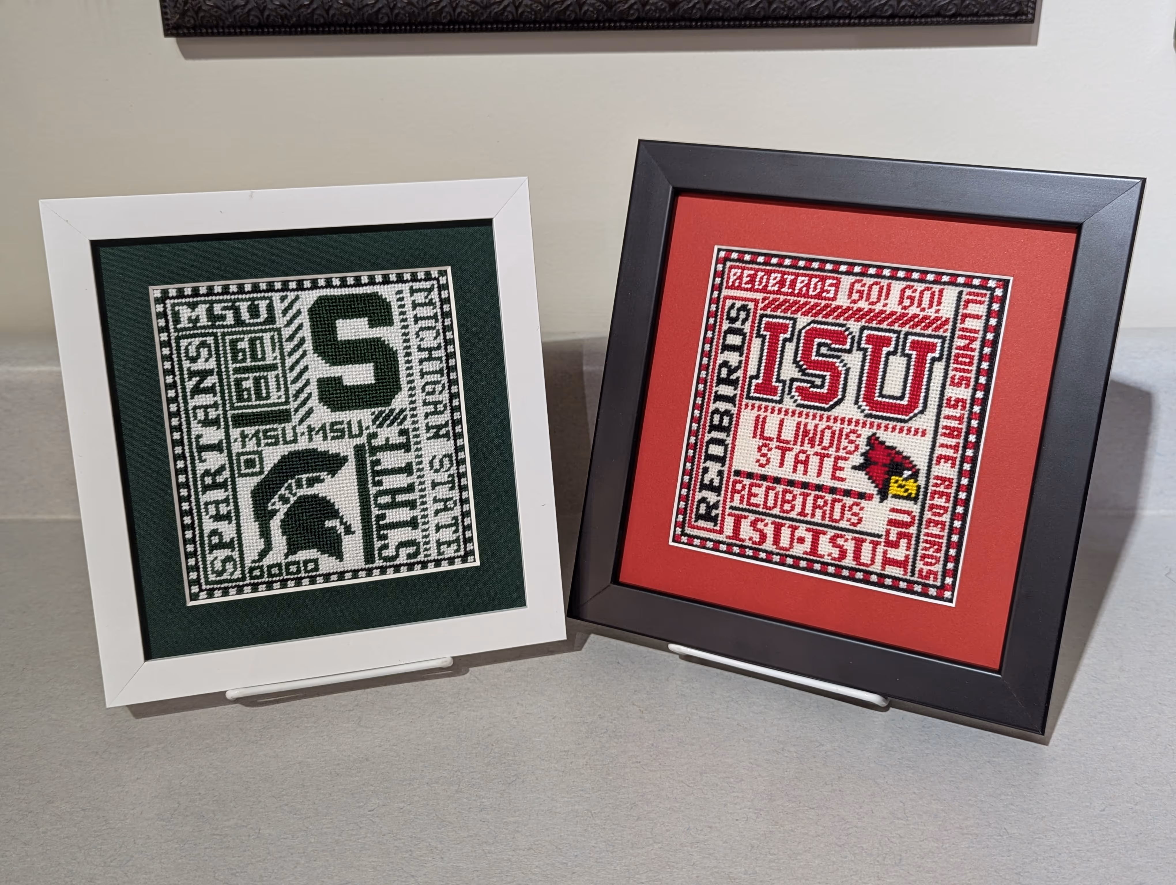 Two framed cross-stitched designs featuring US university themes: Michigan State Spartans in green and white, Illinois State Redbirds in red, black, and white.