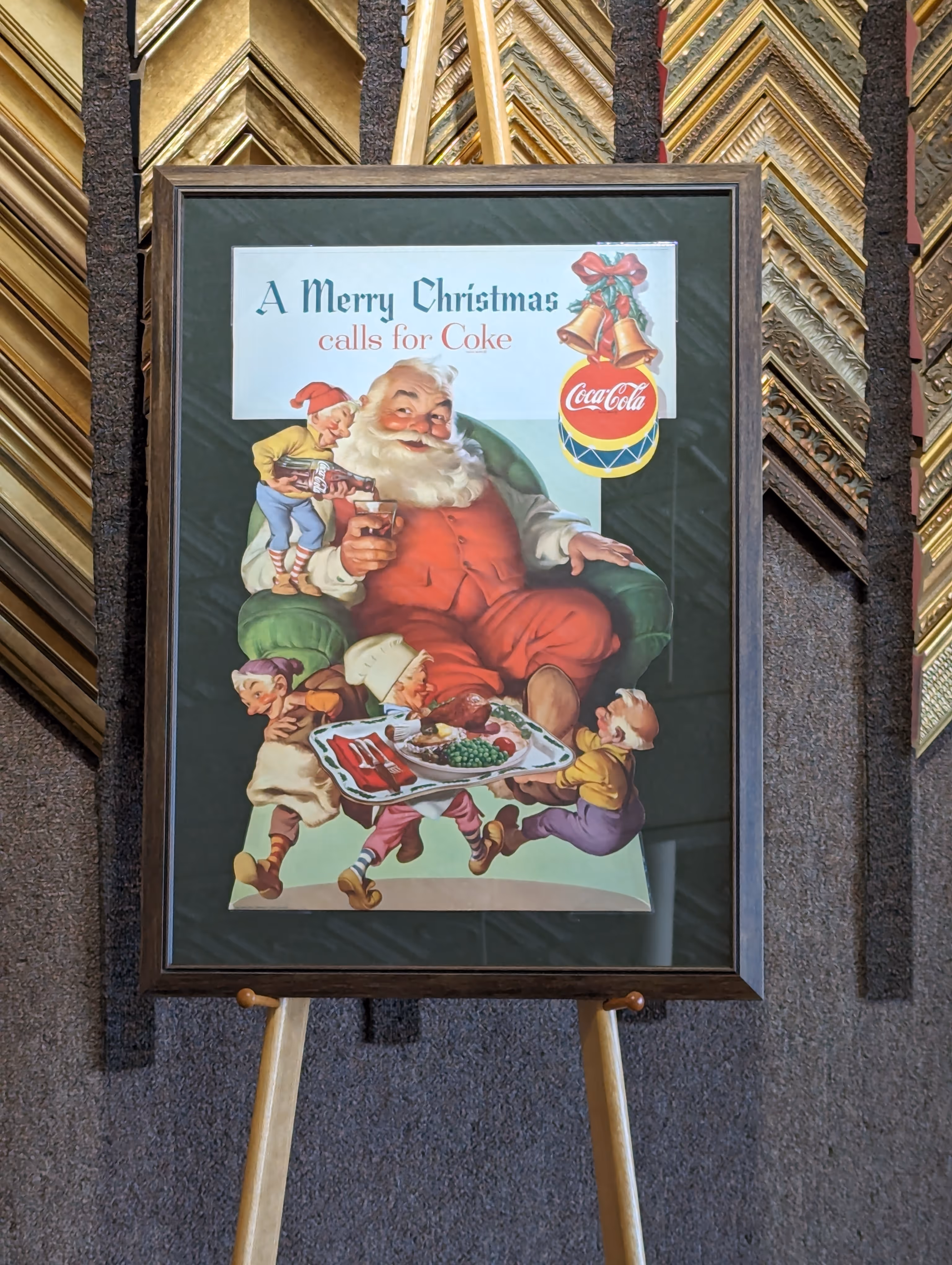 Framed vintage Coca-Cola Christmas poster showing Santa Claus in a red suit sitting in a green armchair, with elves bringing him a tray of food and one elf pouring a bottle of Coca-Cola into his glass.