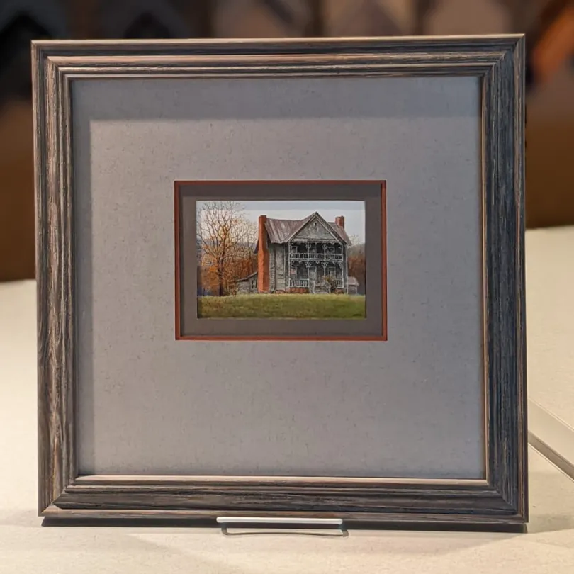Framed miniature painting of an old, two-story rustic house with red brick chimneys, surrounded by autumn trees and grass.