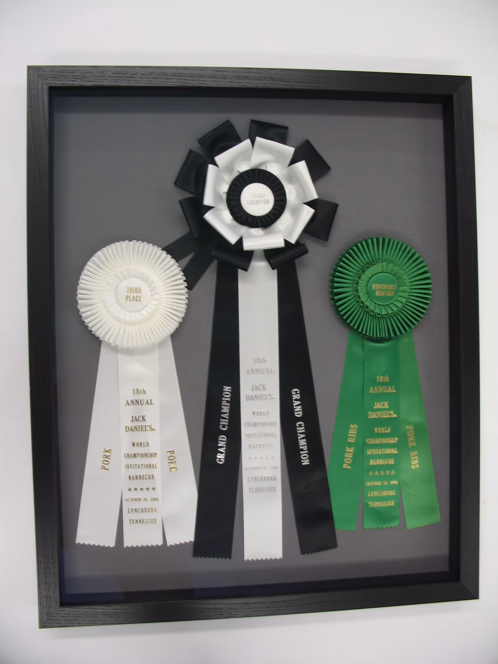 Three award ribbons in a black frame from the 18th Annual Jack Daniel's World Championship Invitational Barbecue in Lynchburg, Tennessee, including third place, grand champion, and honorable mention.
