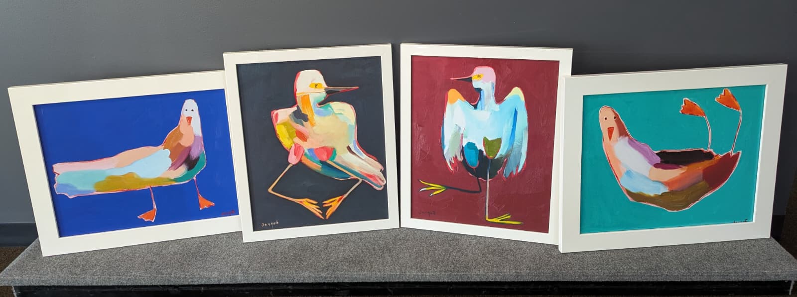 Four colorful abstract bird paintings in white frames displayed side by side on a gray surface against a dark background.