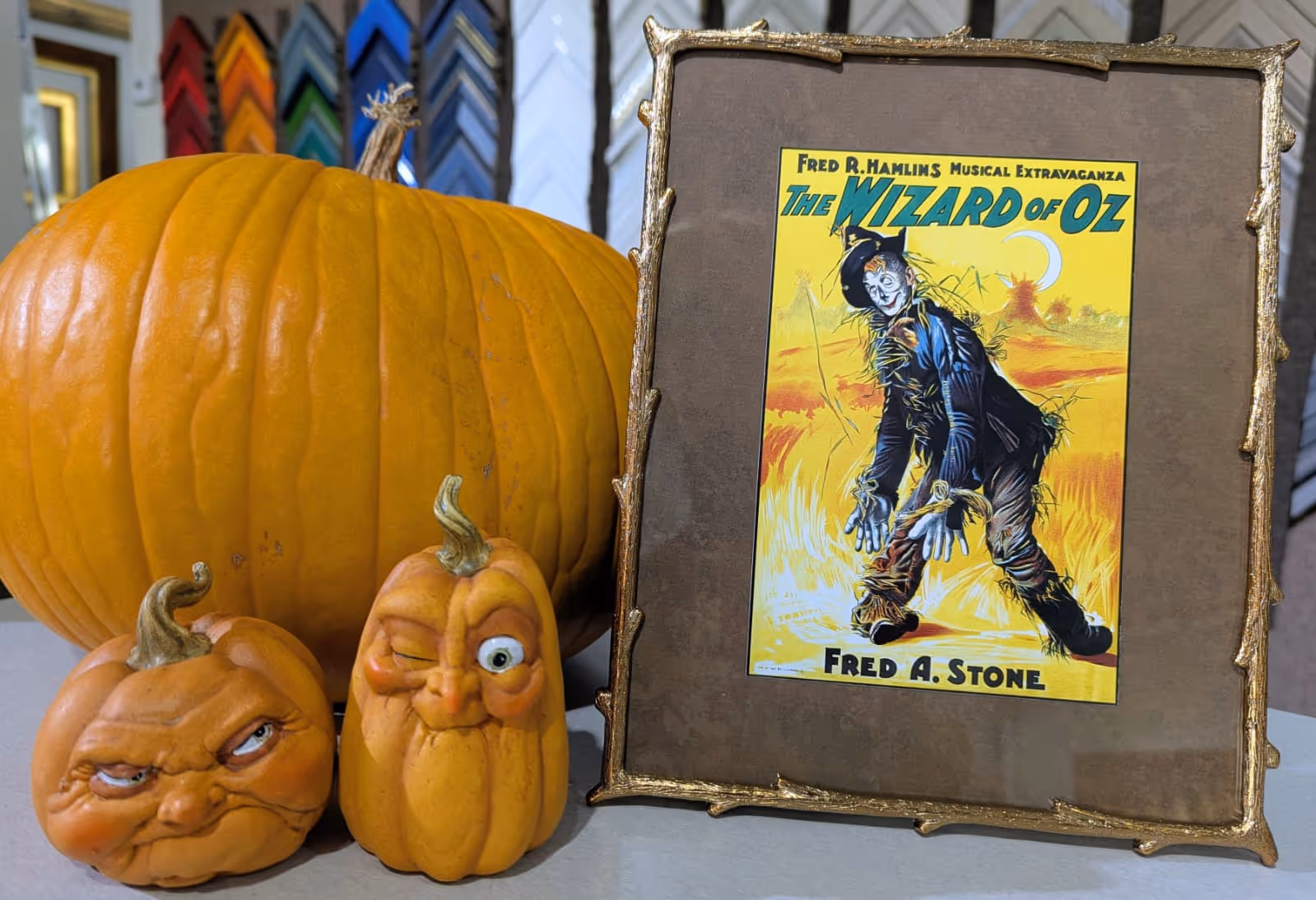 Three pumpkins including two small ones with carved faces and a framed poster of the Wizard of Oz featuring a scarecrow on a yellow background.