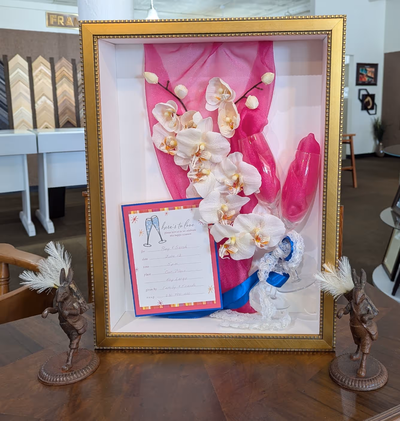 Shadow box frame with pink fabric, white orchids, two champagne glasses with pink napkins, and a wedding invitation card inside, displayed on a wooden table between two rabbit figurines.