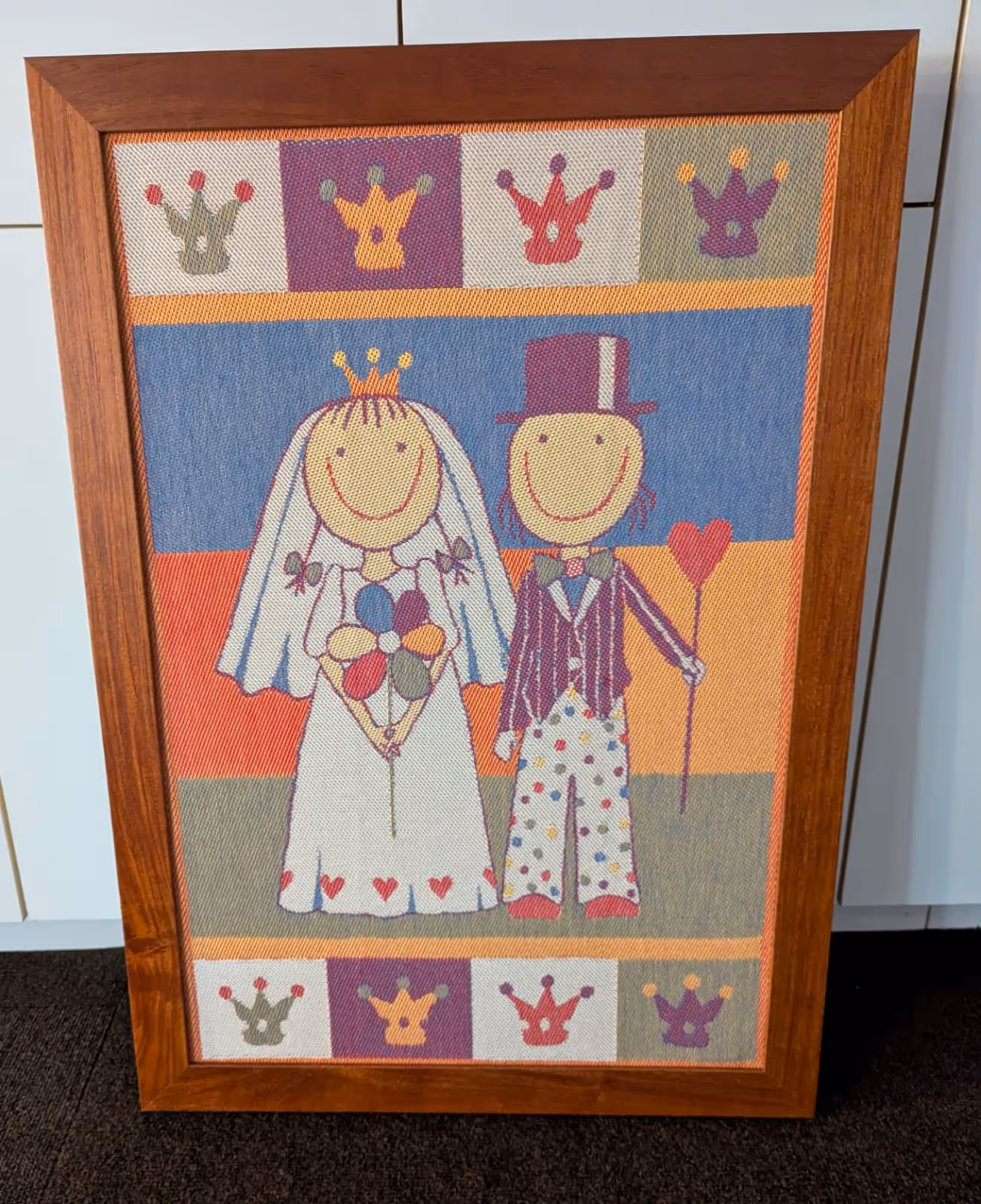 Framed textile artwork showing a smiling bride with a crown and bouquet, and a groom in a top hat holding a heart-shaped staff, surrounded by colorful crowns.