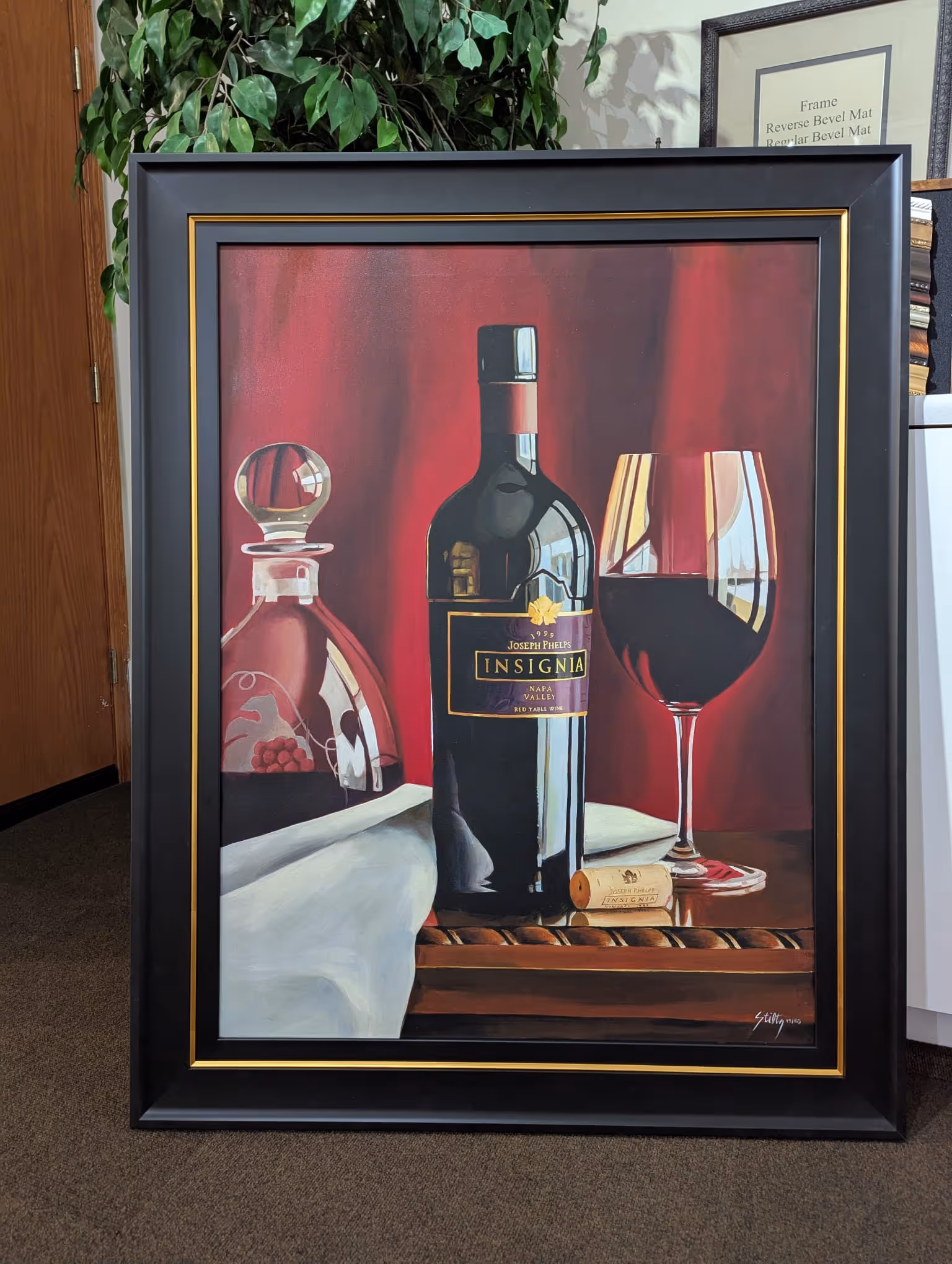 Framed painting depicting a bottle of Joseph Phelps Insignia red wine, a wine glass filled with red wine, a cork, and a glass decanter against a red background.