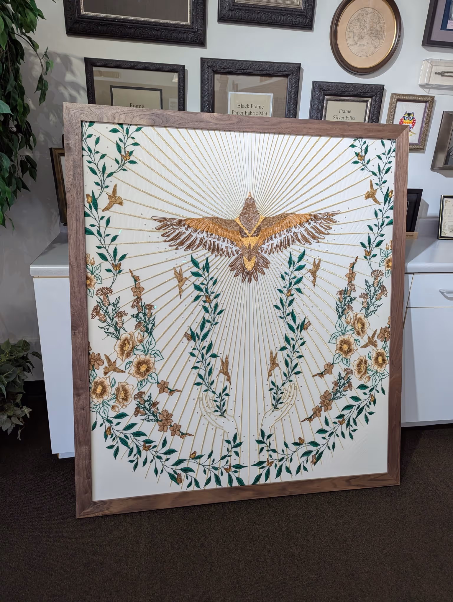 Framed embroidery artwork with a large bird spreading its wings surrounded by flowers, leaves, smaller birds, and two hands reaching upward.