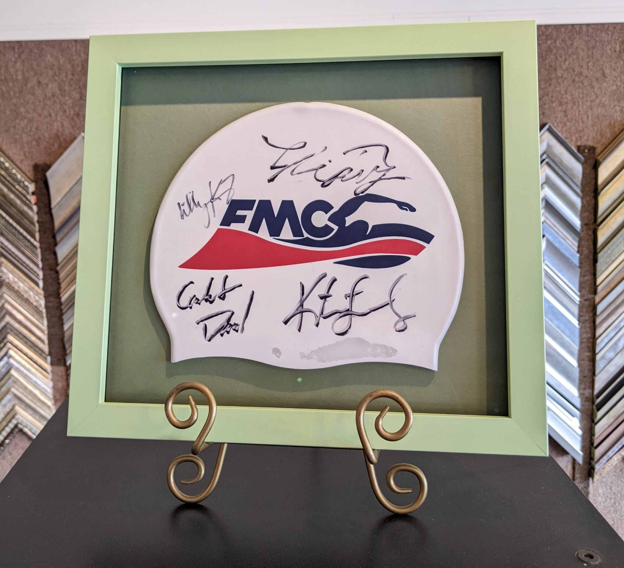 White swim cap with FMCS logo and multiple signatures, displayed in a light green frame on a decorative stand.