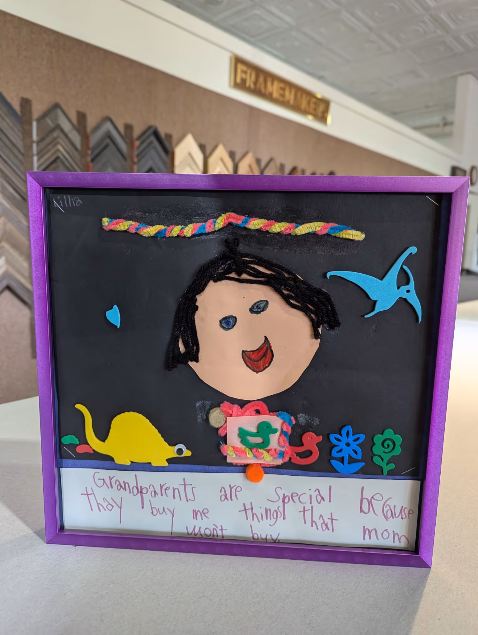 Framed kids artwork featuring a smiling face with black yarn hair, colorful craft shapes including a yellow dinosaur, blue bird, red duck, and flowers, with a handwritten note about grandparents in purple marker.