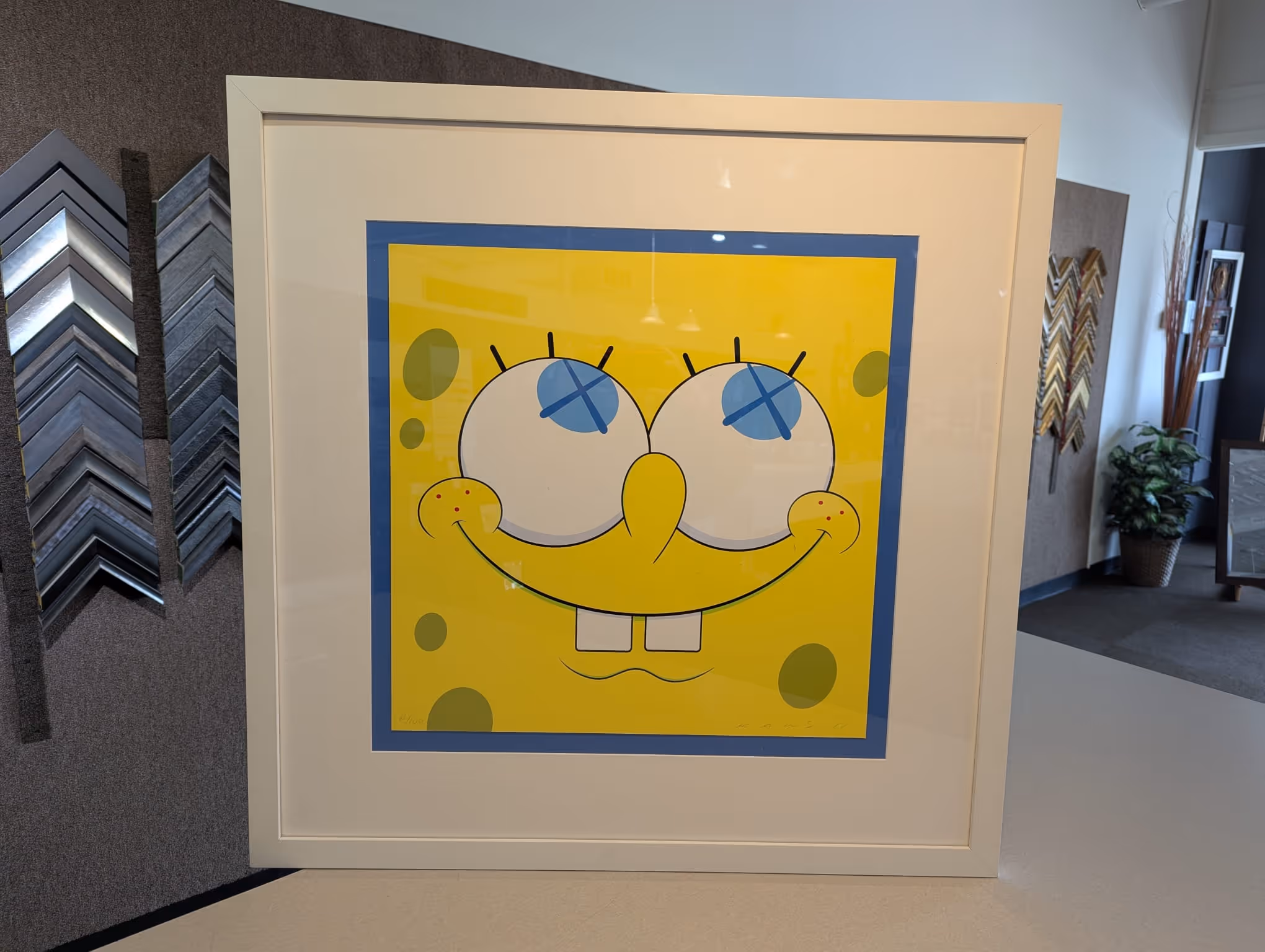 Framed artwork of a yellow cartoon character with crossed-out blue eyes and a big smile, resembling SpongeBob SquarePants.