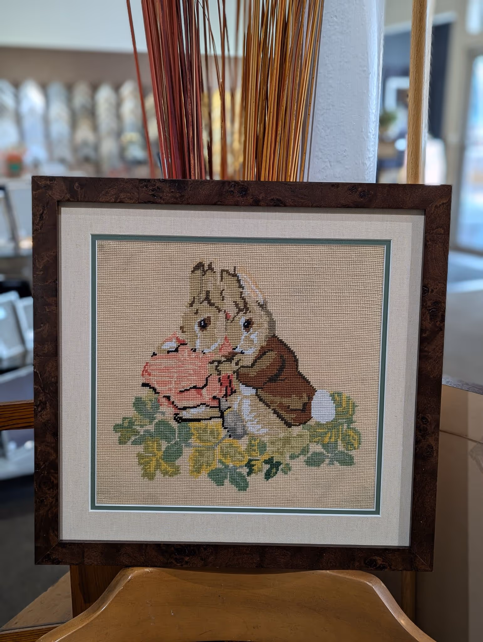 Framed cross-stitch artwork of two rabbits, one wearing a pink dress and the other a brown jacket, sitting among green and yellow leaves.