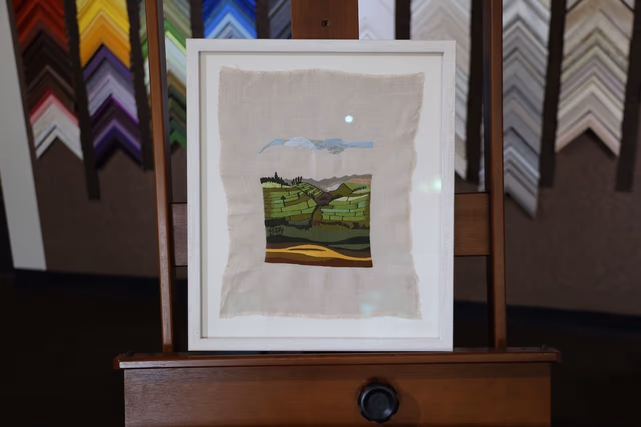 Framed textile artwork depicting a green landscape with hills and a blue sky, displayed on a wooden easel with various colorful picture frame samples in the background.