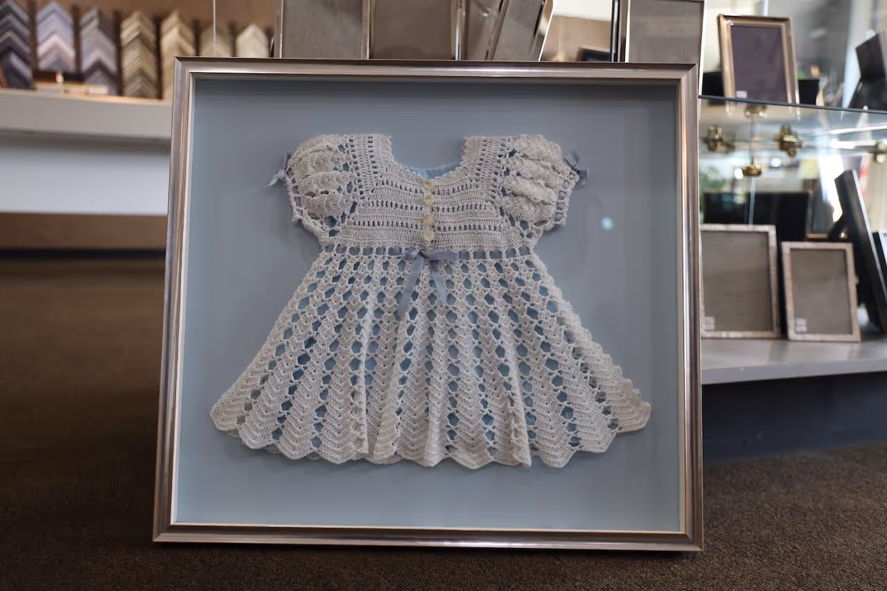 Framed vintage white crocheted baby dress with puffed sleeves displayed on a blue background.