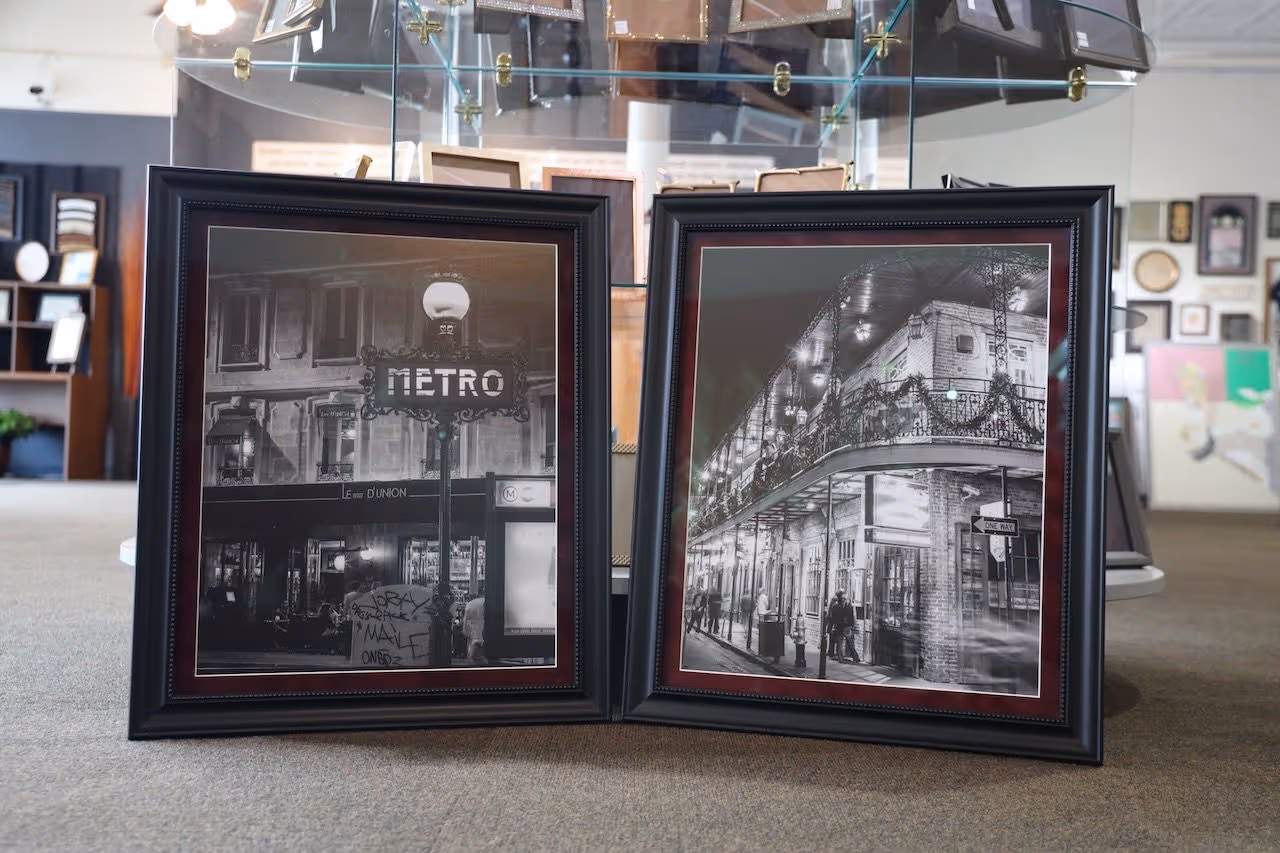 Two framed black and white photographs leaning against a counter, depicting a Metro sign and a historic building with wrought-iron balconies at night.
