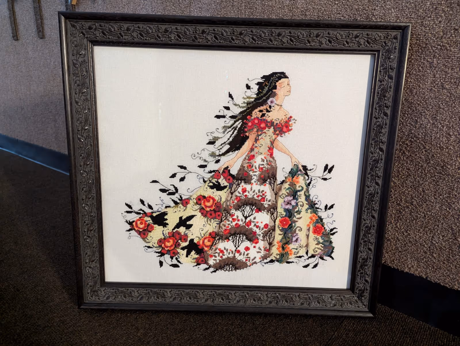 Framed cross-stitch artwork of a woman in a floral dress holding up parts of the skirt with flowing black hair and closed eyes.