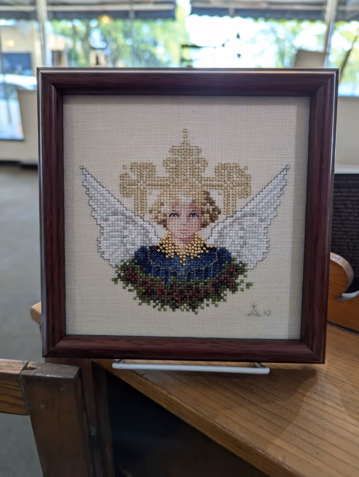 Framed cross-stitch artwork of an angel with blonde hair, white wings, and a golden collar, surrounded by a green and red floral garland.