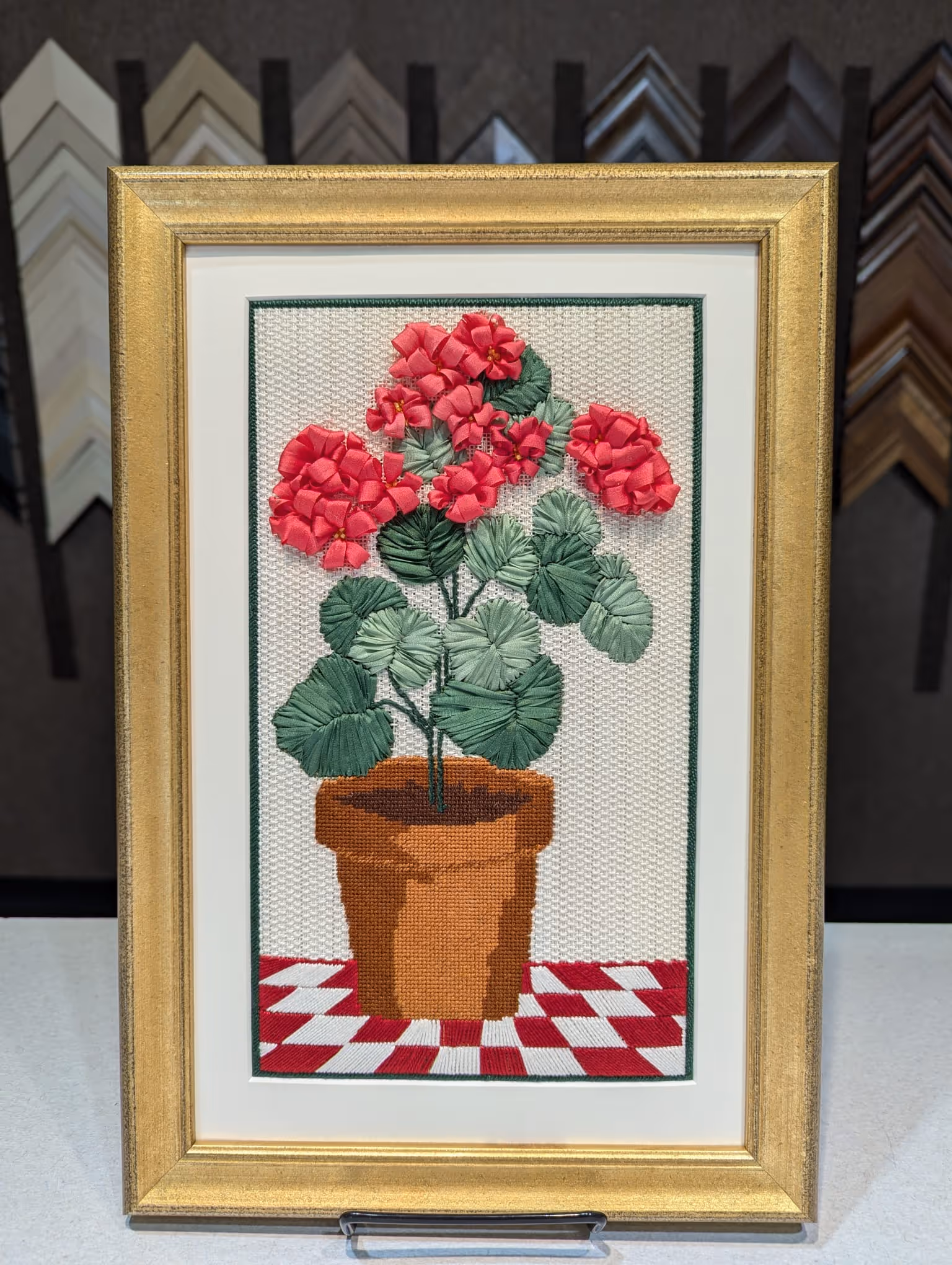 Framed needlepoint embroidery of a terracotta flower pot with pink flowers and green leaves on a red and white checkered surface.