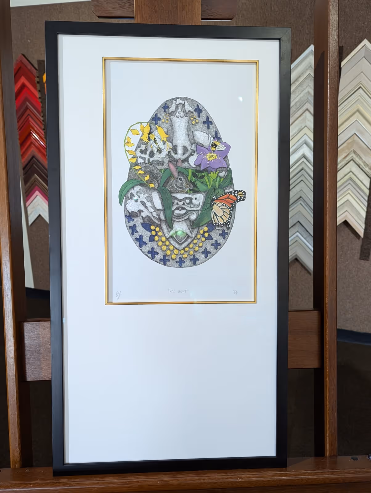 Framed artwork titled 'Egg Hunt' featuring a decorative egg with a rabbit, yellow and purple flowers, a bee, and a monarch butterfly.