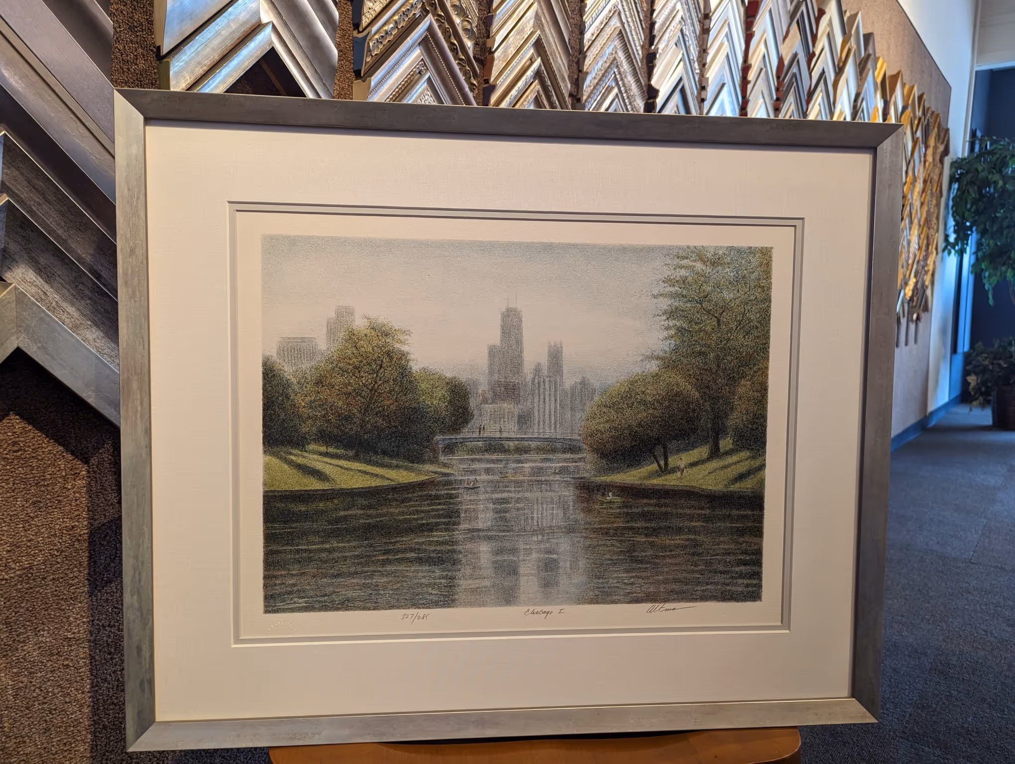 Framed artwork depicting a river with the Chicago skyline and trees on both sides under a cloudy sky.