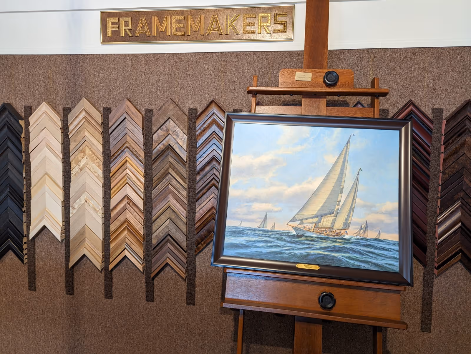 Framed painting of sailboats on the ocean displayed on a wooden easel, with various frame samples on a wall behind.