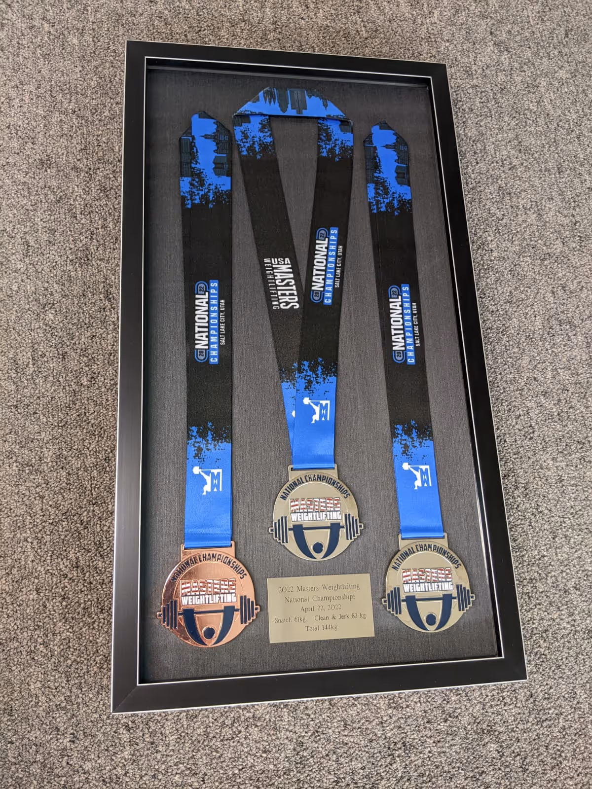 Framed display of three Masters Weightlifting National Championships medals with blue and black ribbons, and a plaque detailing the 2022 event results.