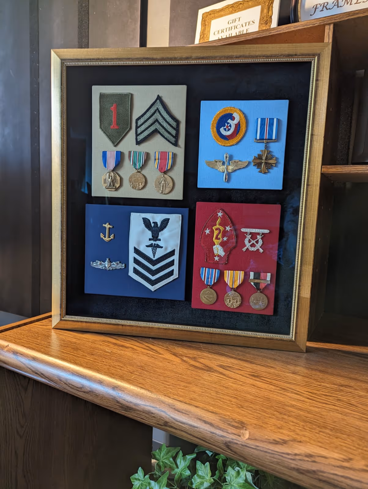 Framed display of four panels featuring various military patches, ribbons, and medals on a wooden surface.