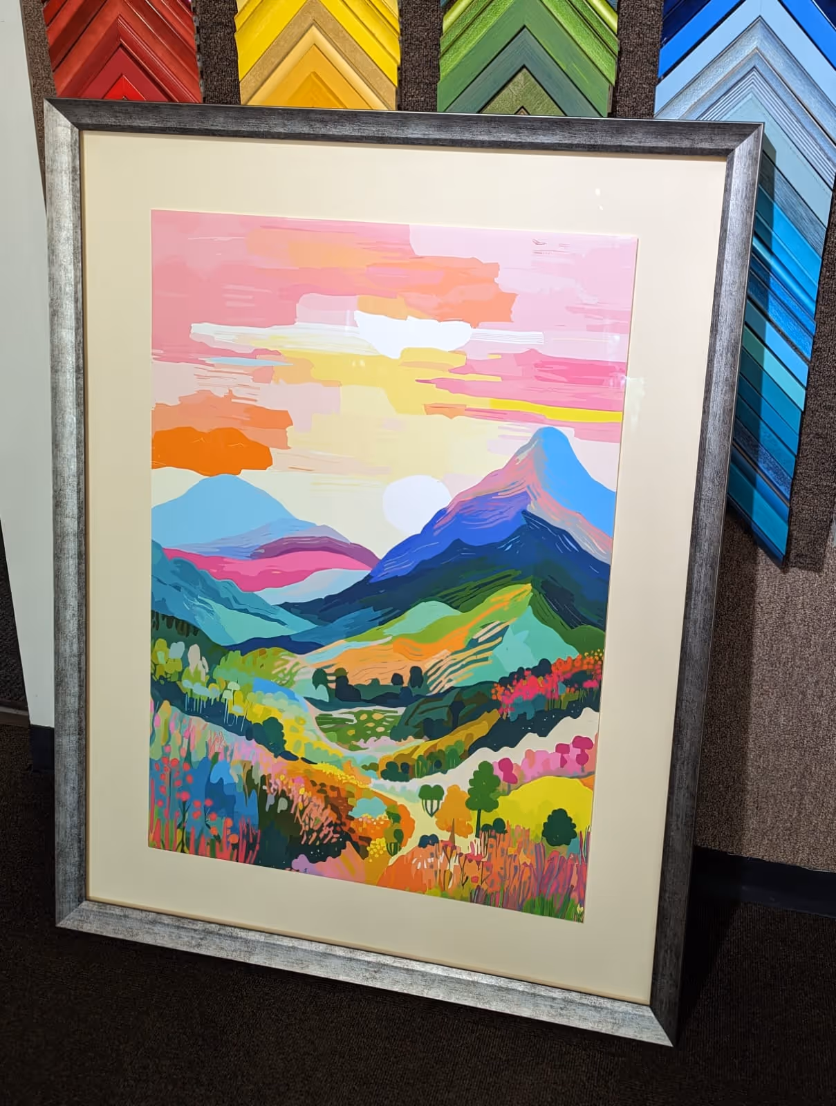 Framed colorful painting of a vibrant mountain landscape with pink sky, blue mountains, and multicolored wildflowers.