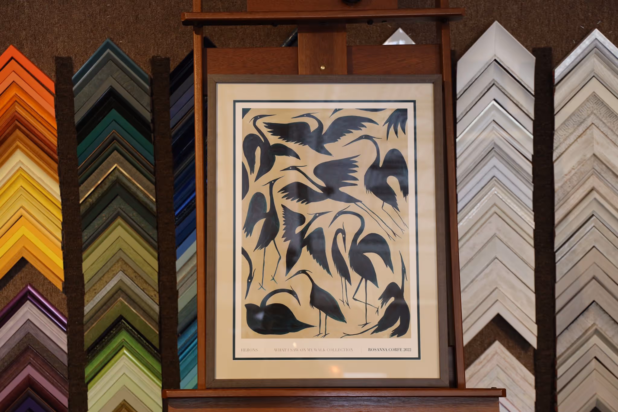Framed artwork depicting multiple black silhouette herons in various poses on a beige background, displayed on a wooden easel with colorful picture frame samples behind it.