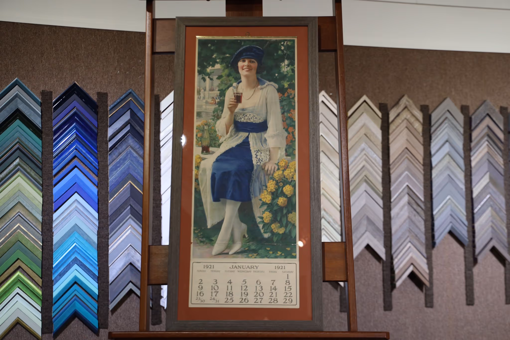 Framed vintage 1921 calendar featuring a woman in a blue dress and hat holding a glass of Coca-Cola, surrounded by flowers.