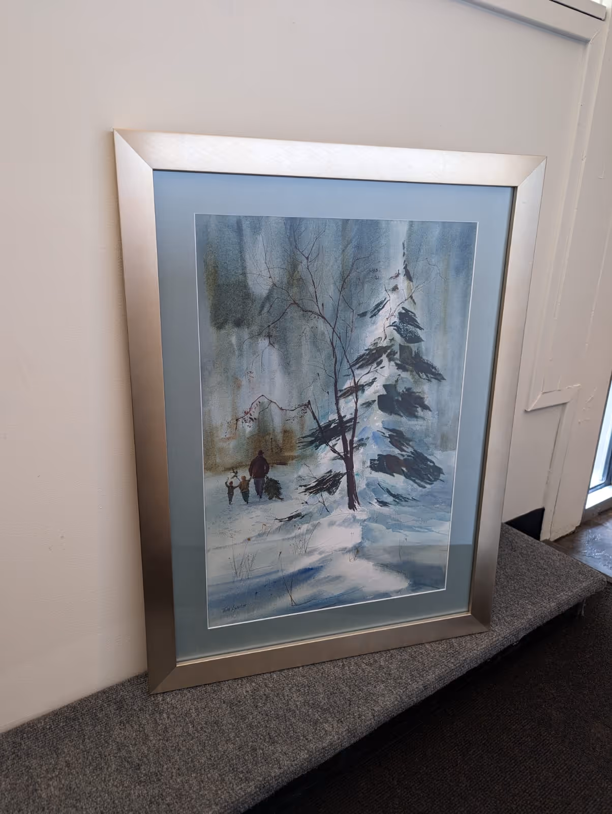 Framed painting depicting a snowy winter scene with a large snow-covered tree and a person walking with two children carrying a Christmas tree.