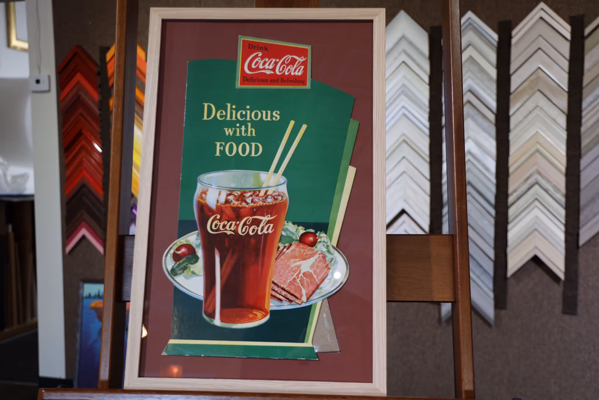 Framed vintage Coca-Cola advertisement showing a glass of Coca-Cola with two straws and a plate of sliced meat and vegetables, with text 'Delicious with Food'.